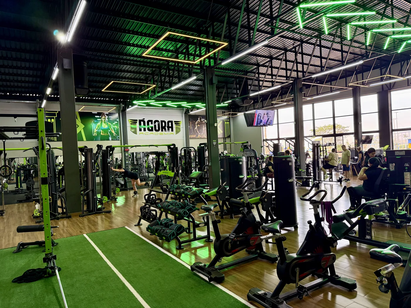 Área de musculação da Ágora com aparelhos novos, bikes de spinning e faixa de grama sintética, sob iluminação verde