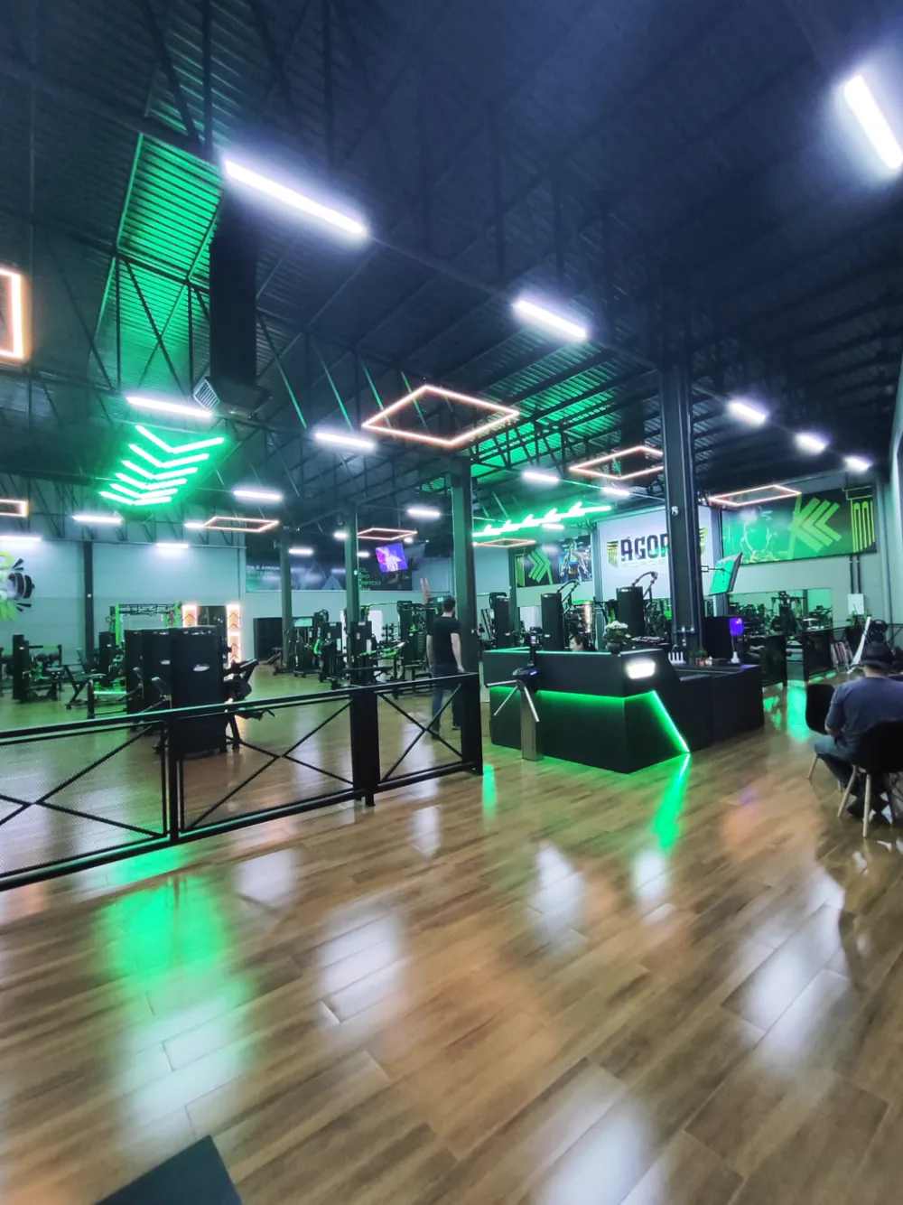 Recepção da Ágora com catraca de acesso e piso amadeirado, com o salão de musculação ao fundo